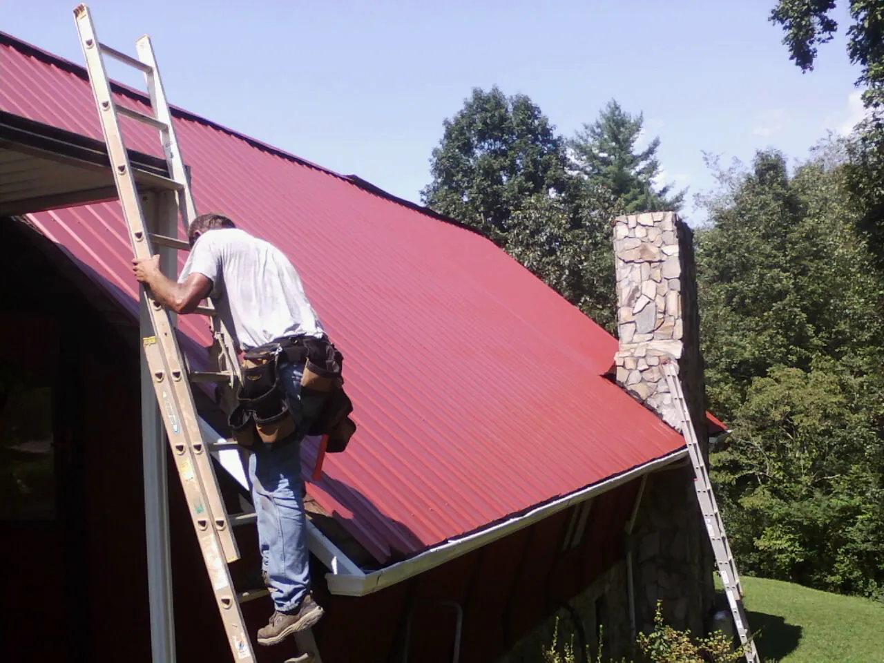 Roofer on a ladder beside a red metal roof and stone chimney — Soffit & Fascia Repair by Roofing Brothers in North Ridgeville